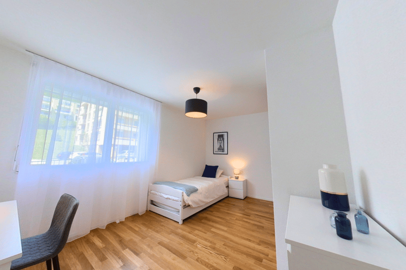 A single bed, nightstand with lamp, nightstand with picture, window with curtains, chair, and wooden floor.