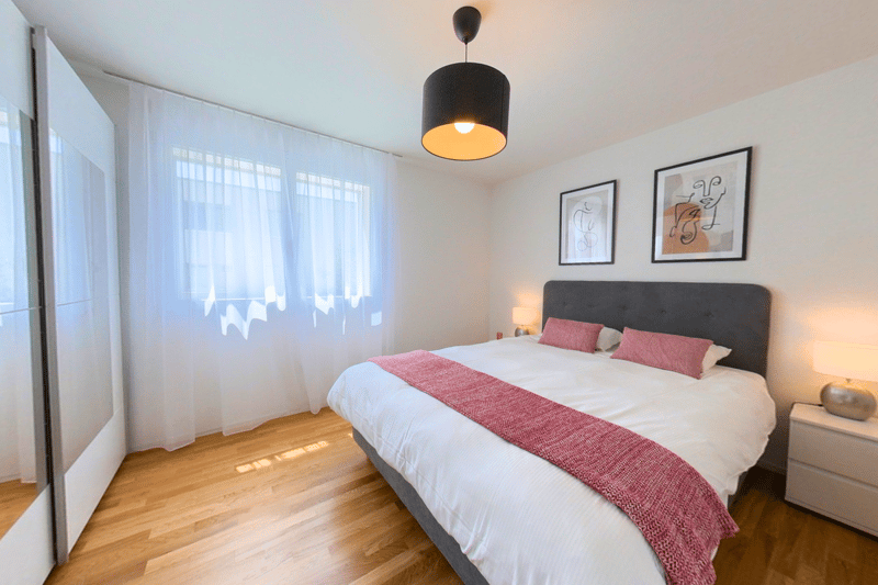 Bedroom with wooden floor, two nightstands with lamps, wall decor, bed with pillows, and white curtains
