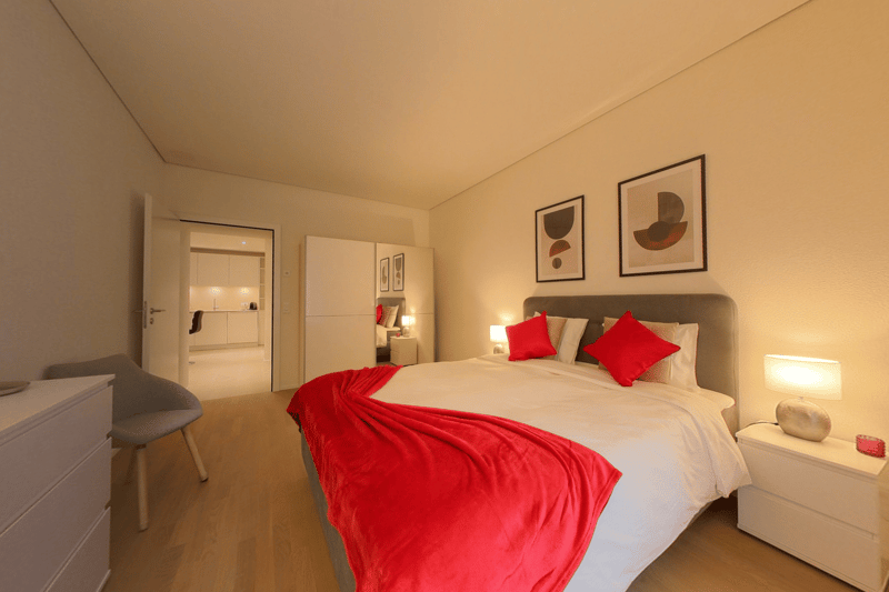 Bedroom with double bed, red pillows, grey headboard, bedside table with lamp, picture frames on the wall, gray chair, white furniture, hardwood floor