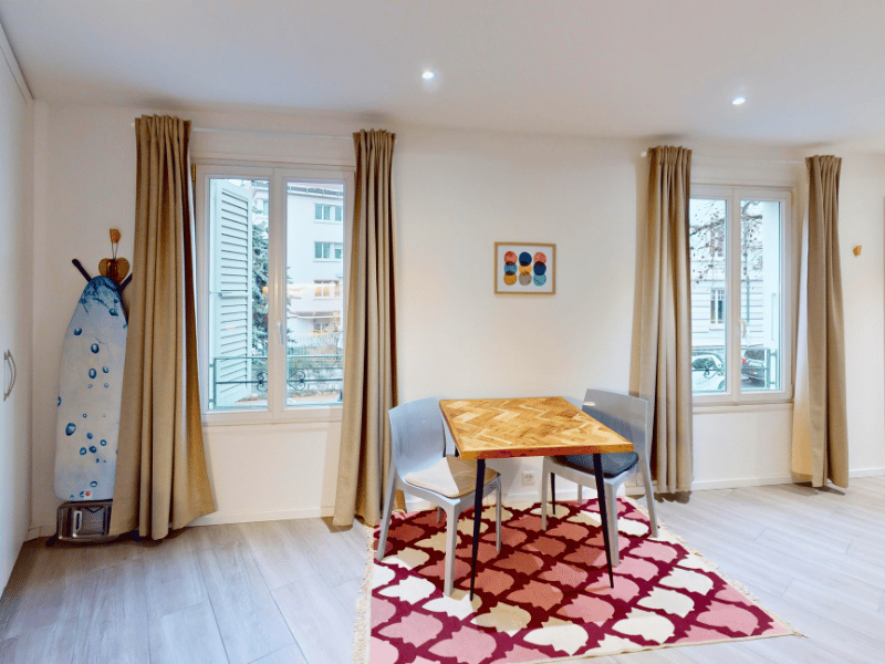A living room with a small dining table, rug, and two chairs. Windows with curtain.