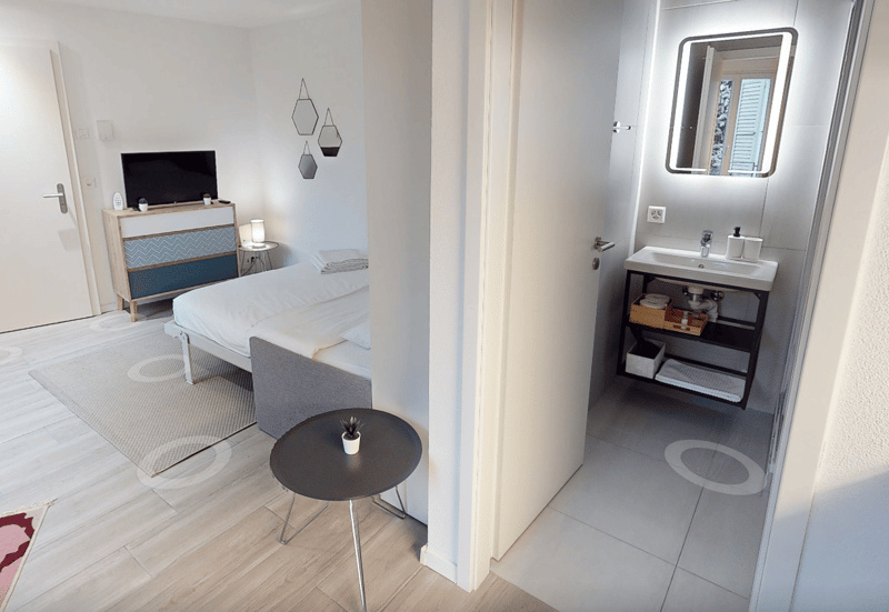 Spacious bedroom with comfortable bed, nightstand, desk, and large windows. Modern bathroom with white sink and mirror. Stylish room decor and neutral color scheme.