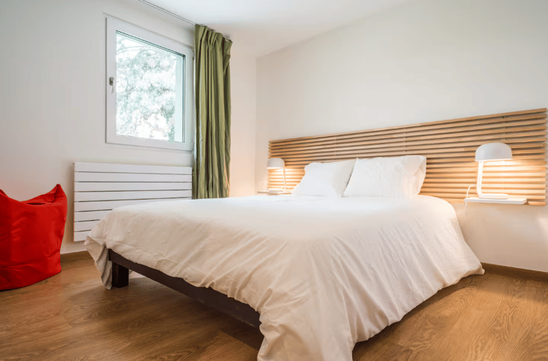Bedroom with wooden bed, bedside tables with lamps, radiator, white bedding, window, curtain