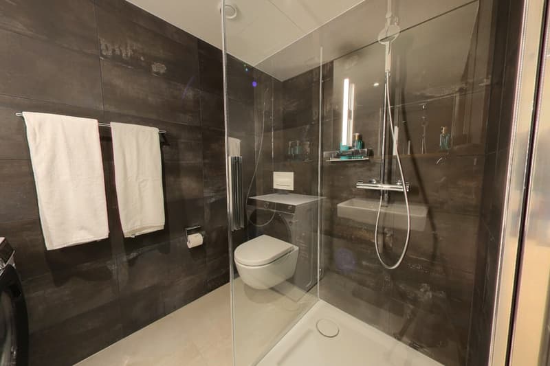 Modern bathroom with a shower, white tiled floor, two towels hanging, a toilet, sink, and a washing machine inside the shower.