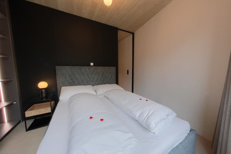Bedroom with double bed, two white pillows, black wall, wooden ceiling, wooden shelves, wooden nightstand with lamp.