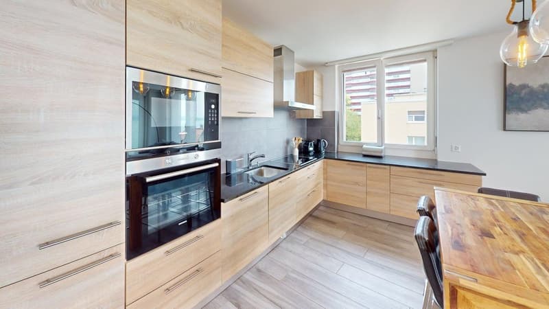 Wooden cabinets, black countertops, stainless steel appliances, wooden table, two chairs, bright window, pendant lights.