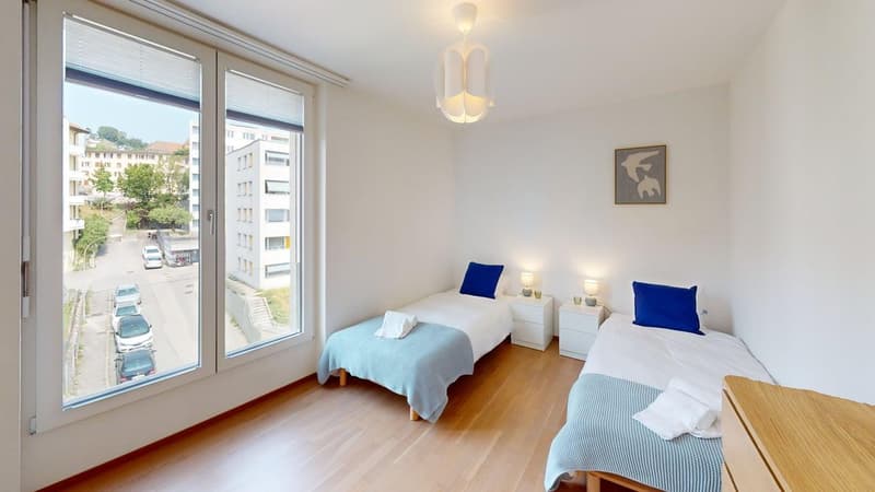 Two single beds, white walls, blue pillows, bedside tables, lamps, wooden floor, large window, towel on bed, large painting on wall.