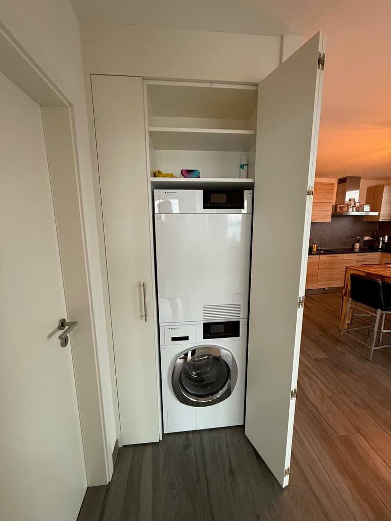 The image shows a utility room with a washing machine and a dishwasher installed in a built-in cabinet. The room has white walls and a wooden floor.