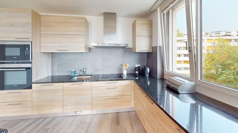 Modern kitchen with wooden cabinets, granite countertop, stainless steel appliances, a sink, faucet, window with a view of buildings and trees outside, black countertop, toaster on the counter, a black and silver toaster, and a kettle.