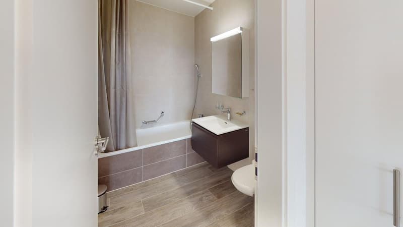 Bathtub, white walls, tiled flooring, wall-mounted sink, shower head, toilet, trash bin, mirror, lighting above mirror