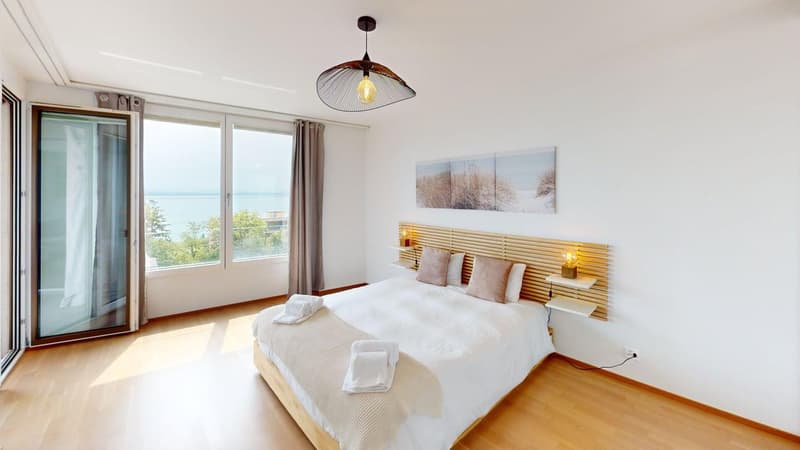 wooden floor, white walls, large windows with sliding doors, wooden bed with white linens, wooden headboard with built-in shelves, bedside lamps, wall art, balcony, sea view
