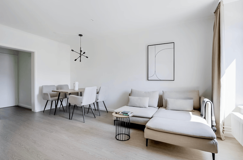 Spacious open-plan living and dining area with white walls, hardwood flooring, a sectional sofa, a dining table with chairs, and a modern light fixture. There is also a framed artwork on the wall and a small side table.