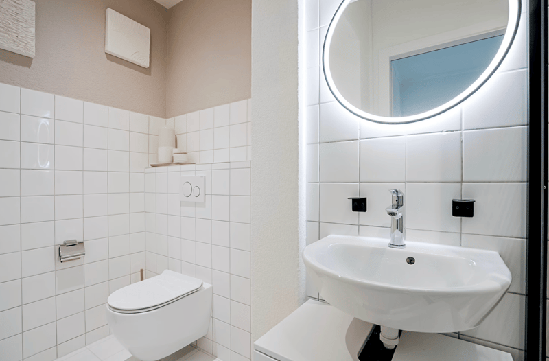 White tiles, modern fixtures, toilet, sink, round mirror, minimalistic