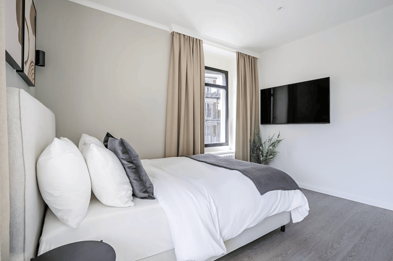 Modern styled bedroom, queen size bed with white sheets and a gray blanket, grey bed headboard, two lamps, potted plant, mounted TV, white walls, curtained window, gray floors