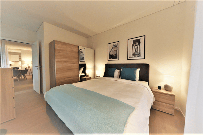 Spacious bedroom with a large bed, wooden wardrobe, and framed artwork on the walls. The room has a neutral color scheme with beige walls and a blue bedspread. There appears to be a balcony or terrace visible through the doorway.