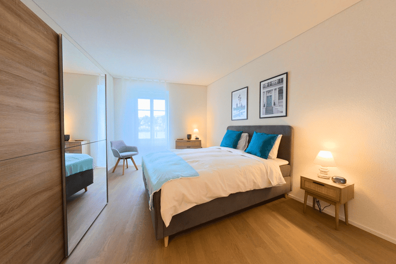 Bed with white sheets and blue blankets, two nightstands with lamps, wooden bedside table, mirror, chair, framed pictures, window with white curtains.