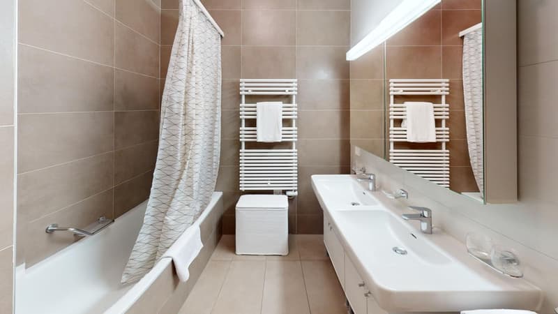 Spacious bathroom with beige tile walls, white bathtub, double vanity sink, and towel warmers