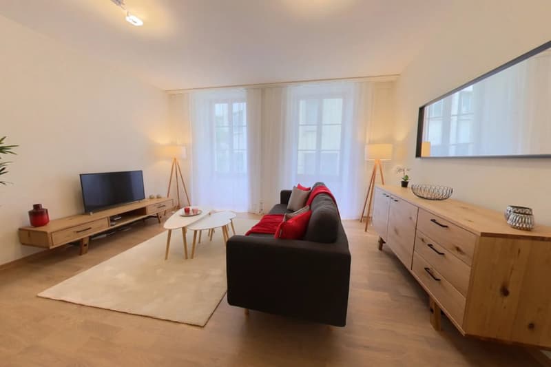 Spacious living room with large windows, hardwood floors, a black sofa, a wooden TV stand, and a coffee table. The room has a cozy and modern feel with warm lighting and decorative elements.