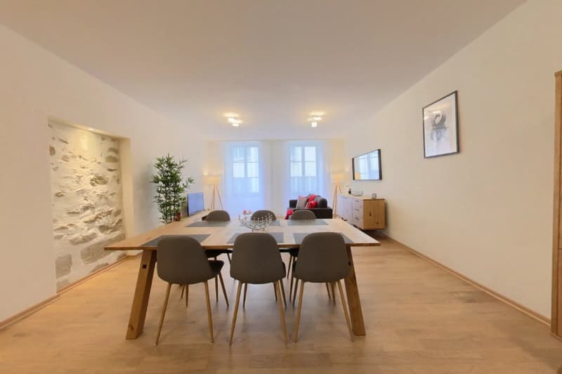 Spacious dining area with a large wooden table and six gray chairs. The room has white walls, large windows, and a stone accent wall. There is a potted plant and some artwork on the walls.