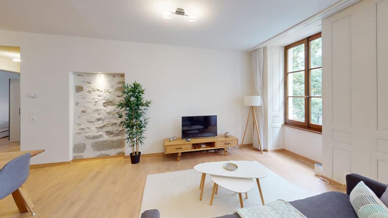 Spacious living room with large windows, hardwood floors, white walls, wooden TV stand, coffee table, and a potted plant.