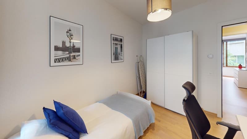 Bedroom with hardwood floor, white walls, large window, wardrobe, bed with blue bedding, and framed artwork on the walls.