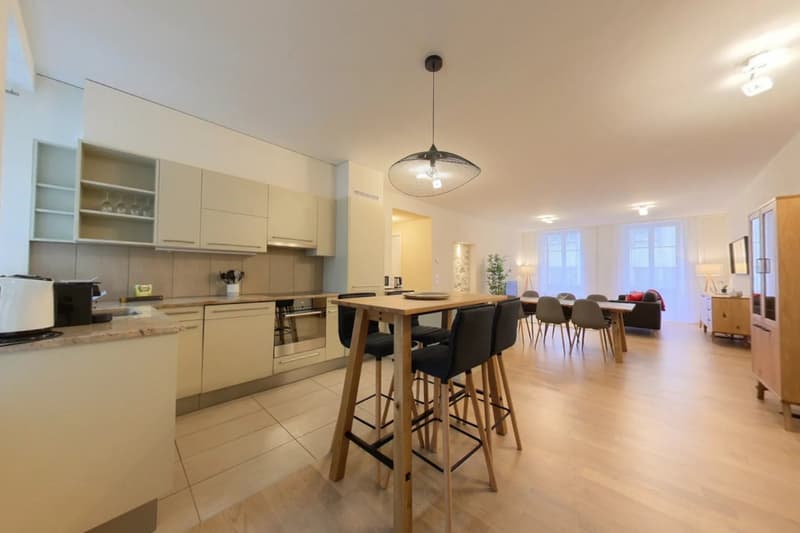 The image shows an open concept kitchen and dining area with a modern, minimalist design. The kitchen features light-colored cabinets, a built-in dishwasher, and a tiled backsplash. The dining area has a large wooden table with black bar stools, and the s