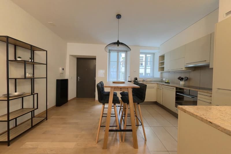 Open concept kitchen with modern appliances, wooden dining table and chairs, shelving unit, and large windows providing natural light