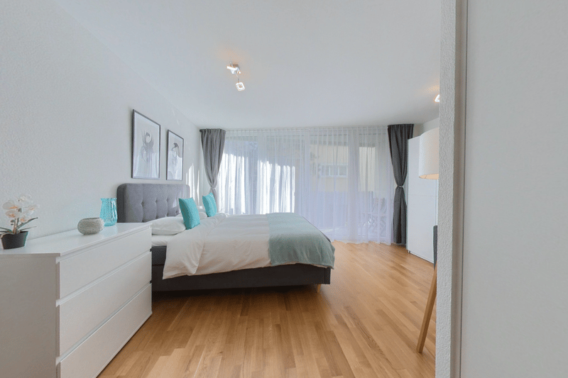 spacious bedroom with large bed, wooden floors, white dresser, decorative pillows, and large windows with curtains