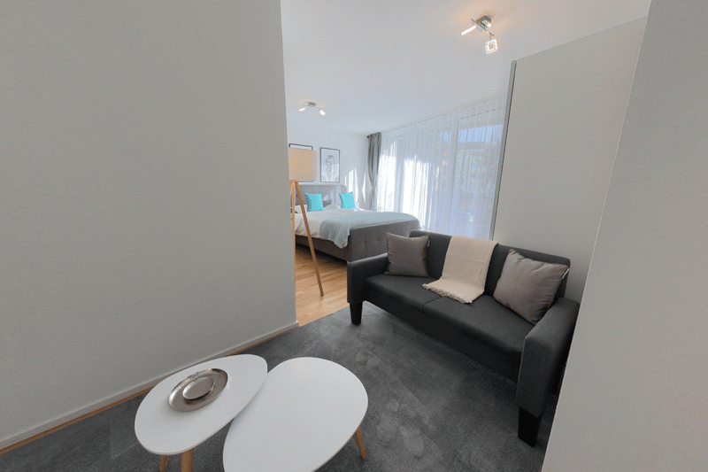 Modern apartment room, gray couch with pillows, white tables, beige rug, open view to bedroom with bed