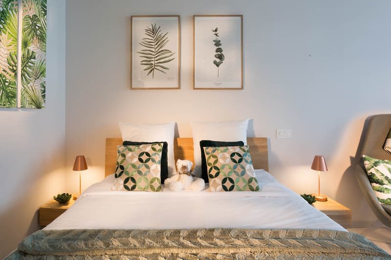 Cozy bedroom with a white bed, patterned throw pillows, and a small stuffed animal. The room features framed botanical prints on the wall and bedside lamps. The large window provides natural light and a view of greenery outside.