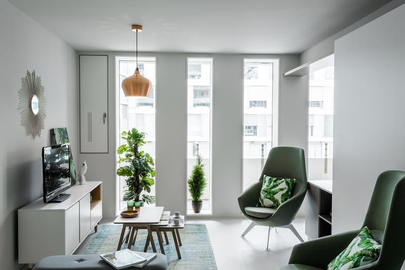 Bright and modern living room with large windows, green accent chairs, TV stand, and potted plants. The room has a minimalist and airy feel with natural light and a view of the surrounding buildings.