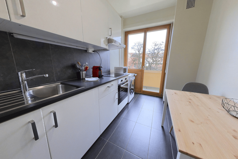 Galley-style kitchen with white cabinets, black countertops, stainless steel sink, dishwasher, and washing machine. Large window providing natural light.