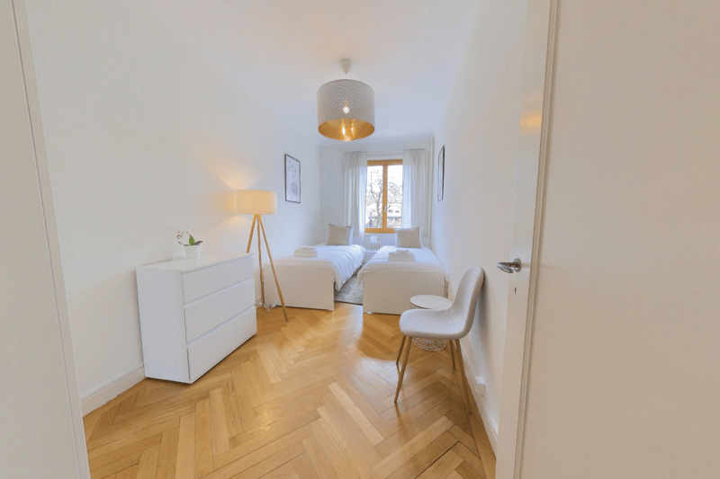 Bright and airy bedroom with hardwood flooring, white walls, two twin beds, a dresser, and a chair. The room has a large window that provides natural light.