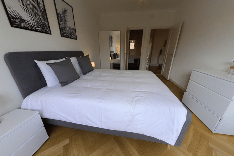 The image shows a bedroom with a large bed with a dark gray headboard and white bedding. There are two framed black and white palm leaf prints on the wall above the bed. The room has hardwood floors and a white dresser on the right side of the room.