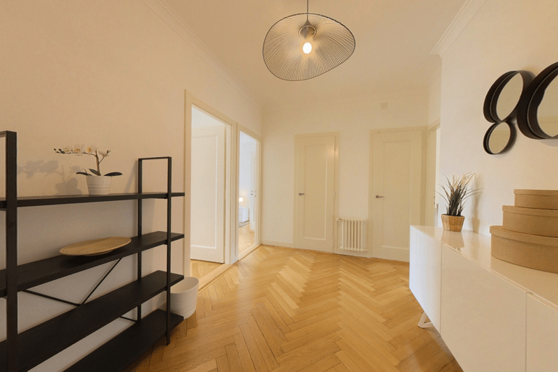 Spacious apartment with hardwood herringbone floors, white walls, black metal shelving unit, potted plant, and a modern pendant light fixture. The room has multiple doors leading to other areas of the apartment.