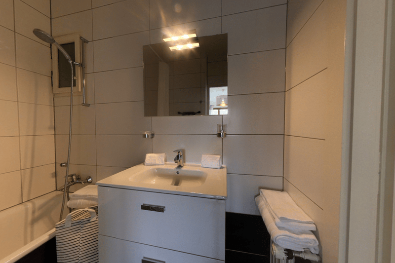 Bathroom with tiled walls, vanity sink with double faucets, mirror, and a storage cabinet below the sink. There is also a shower area with a showerhead.