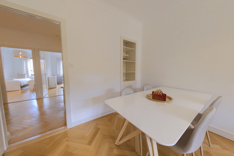 Bright and spacious dining area with a white table and chairs, hardwood flooring, and a built-in shelving unit on the wall. The room has an open layout and leads to an adjacent bedroom area.
