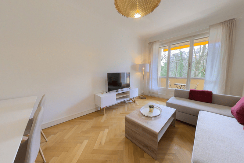 Spacious living room with large windows providing a view, hardwood flooring in a herringbone pattern, white walls, a TV stand, a sofa, and a coffee table with decorative items.