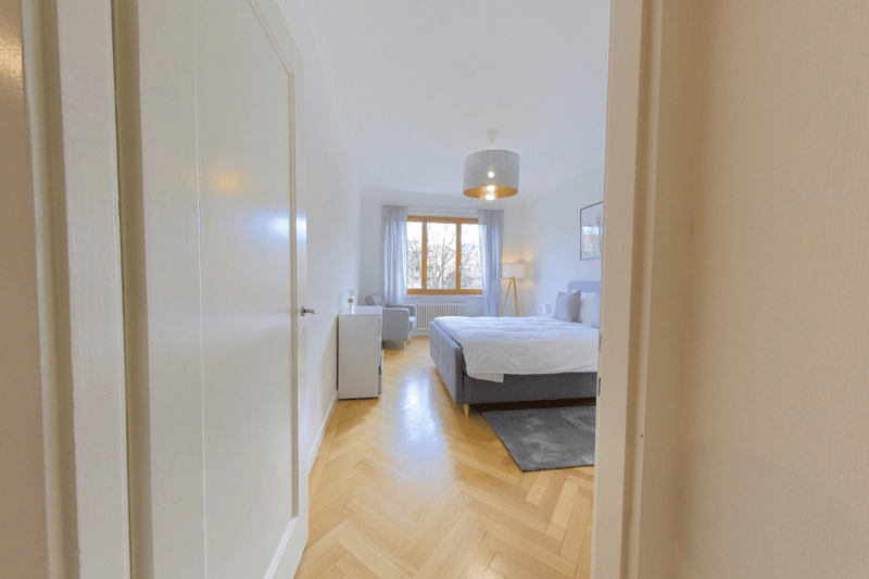 Spacious bedroom with hardwood flooring, large windows, a double bed, a dresser, and a pendant light fixture.