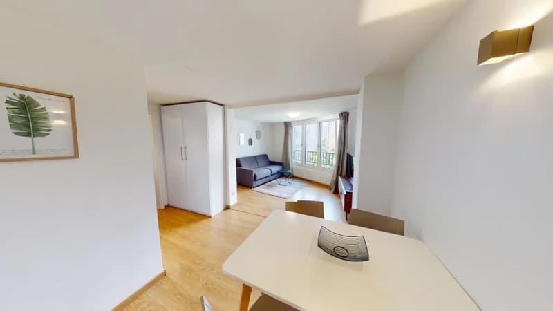Open-concept living space with hardwood floors, white walls, large windows, a sofa, coffee table, and a dining table. There is a balcony or terrace visible through the windows.