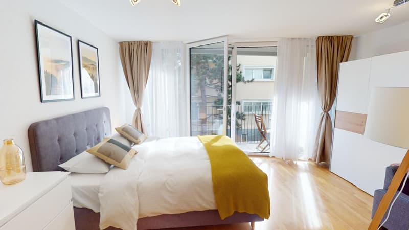 Bedroom with double bed, balcony with outdoor furniture, wooden floor, white walls, beige headboard, two picture frames, white bed linen, glass doors