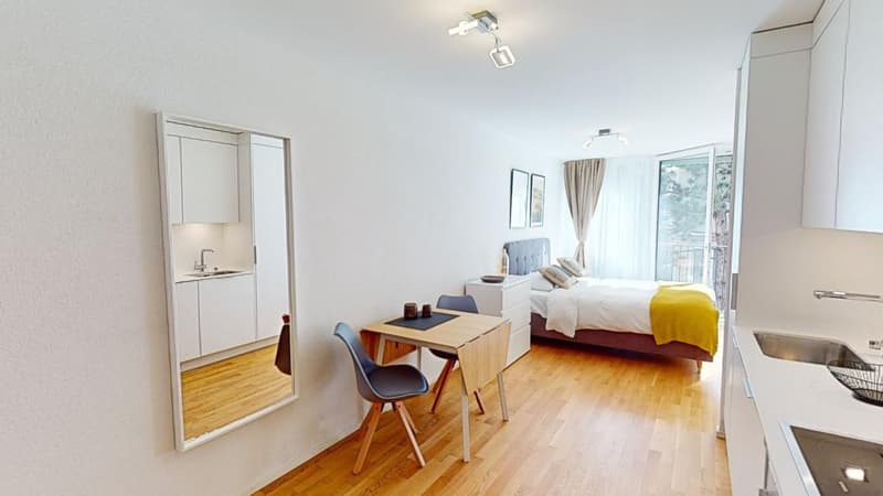 Single room with wooden floor, bed, desk with 2 chairs, kitchen with sink, mirror, lamps, balcony