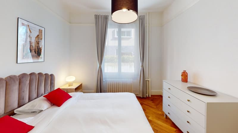 white walls, hardwood floors, large double bed, bedside table with lamp, framed picture, gray curtains, white dresser with plate, vase on dresser