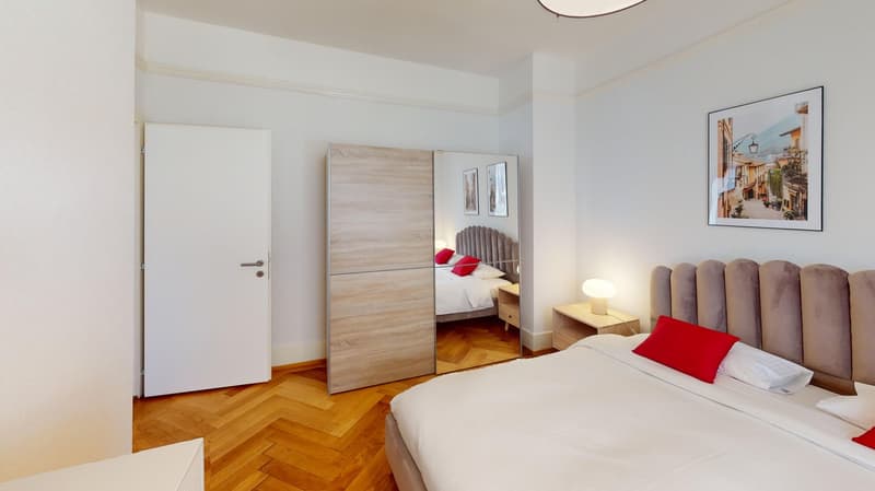 Large bed, wooden floor, white walls, a full-length mirror, a nightstand with a lamp, a wardrobe, and a wall picture.