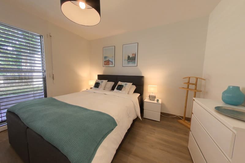 Bedroom with bed, nightstands, lamps, paintings, wooden floor, white walls, large window, wooden stand, white dresser, plate, vase