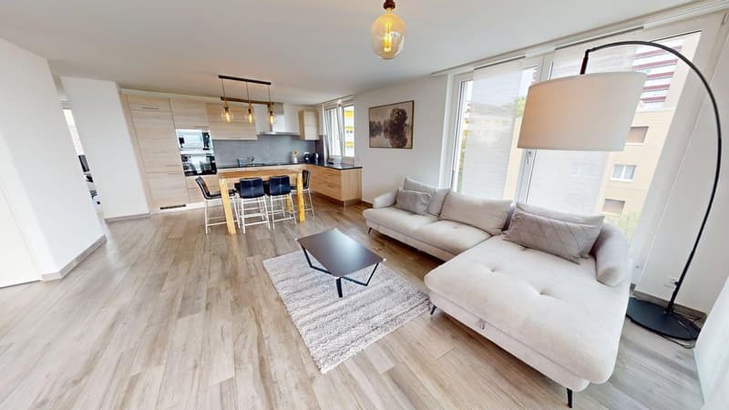 Large living room with a L-shaped white sofa, wooden table and chairs, modern lamp, wooden flooring, white walls, large windows