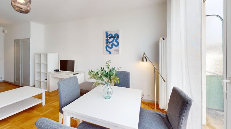 Dining area with a white table, four chairs, a white shelf, a computer, a monitor, a lamp, a radiator, a plant in a vase, and a white curtain.