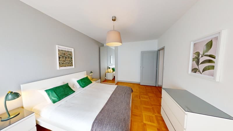 Bedroom with white walls, wooden floor, large bed, nightstands, lamps, mirror, and framed pictures