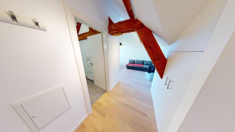 Spacious open-plan living area with hardwood floors, white walls, and exposed wooden beams. Visible is a living room with a blue sofa, and a hallway leading to other rooms.