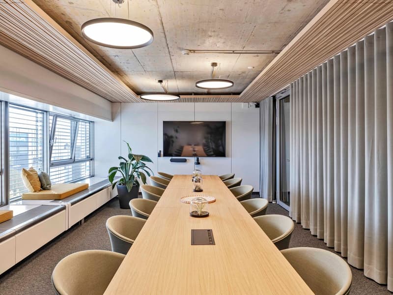 Large wooden conference table with comfortable chairs, modern lighting fixtures, concrete ceiling, large windows, and a TV mounted on the wall.