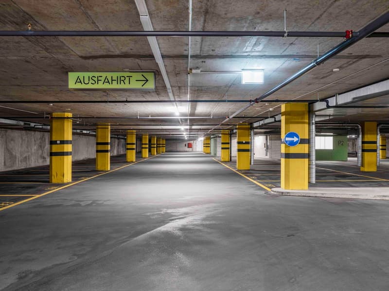 The image shows a large underground parking garage with concrete walls and floors. The space is well-lit, with yellow and black striped columns lining the sides. There is a sign indicating the direction of 'Ausfahrt' or 'Exit'. The overall layout appears